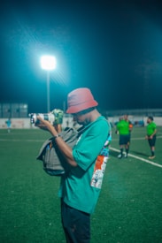 A person wearing a red bucket hat and a green shirt is taking a photograph on a sports field at night. A bright floodlight illuminates the scene. In the background, several other people are visible, wearing green uniforms, suggesting a sports event or practice.
