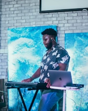 A person is standing at a keyboard synthesizer with a laptop nearby. The background features two paintings with abstract blue designs set against a light-colored brick wall. The individual is wearing a floral-patterned shirt and appears focused on the musical equipment.