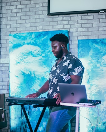 A person is standing at a keyboard synthesizer with a laptop nearby. The background features two paintings with abstract blue designs set against a light-colored brick wall. The individual is wearing a floral-patterned shirt and appears focused on the musical equipment.