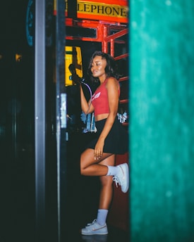A person is standing in a classic red telephone booth, holding the phone to their ear. They are wearing a red crop top, black skirt, and white sneakers with high socks. The setting gives off a vintage and nostalgic feel.