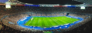 A large football stadium filled with spectators, featuring a well-lit football field in the center surrounded by blue-bordered sections. The stadium is packed with fans, and a match is in progress on the grass field. The stadium lighting highlights the vibrant atmosphere, with large displays showing game information. National flags and banners are visible, adding patriotic elements to the scene.