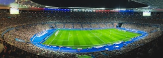 A large football stadium filled with spectators, featuring a well-lit football field in the center surrounded by blue-bordered sections. The stadium is packed with fans, and a match is in progress on the grass field. The stadium lighting highlights the vibrant atmosphere, with large displays showing game information. National flags and banners are visible, adding patriotic elements to the scene.