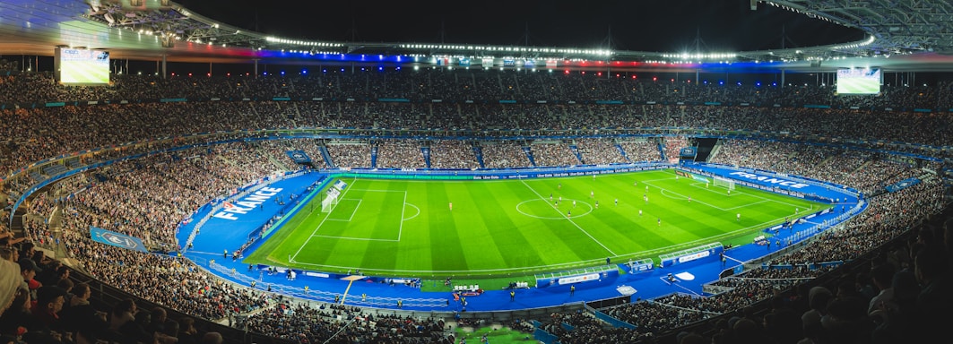 A large football stadium filled with spectators, featuring a well-lit football field in the center surrounded by blue-bordered sections. The stadium is packed with fans, and a match is in progress on the grass field. The stadium lighting highlights the vibrant atmosphere, with large displays showing game information. National flags and banners are visible, adding patriotic elements to the scene.