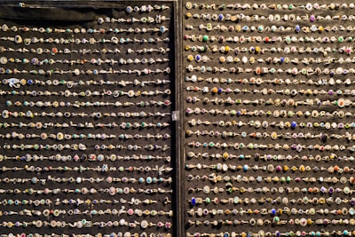 An array of decorative rings is organized in neat horizontal rows against a dark fabric background. Each ring features a variety of gemstones and intricate designs, showcasing a colorful and diverse assortment.