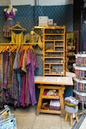 A retail display area showcasing vibrant, patterned clothing items on hangers, including dresses and tops. The wall behind has a decorative geometric pattern and a shelf adorned with fairy lights, while various accessories like jewelry are neatly organized on a wooden table. Below, there's a wooden bench with storage featuring books and a basket.