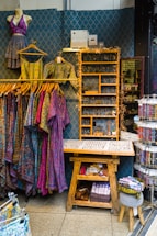A retail display area showcasing vibrant, patterned clothing items on hangers, including dresses and tops. The wall behind has a decorative geometric pattern and a shelf adorned with fairy lights, while various accessories like jewelry are neatly organized on a wooden table. Below, there's a wooden bench with storage featuring books and a basket.