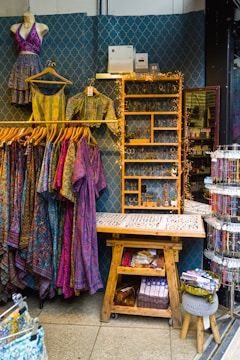 A retail display area showcasing vibrant, patterned clothing items on hangers, including dresses and tops. The wall behind has a decorative geometric pattern and a shelf adorned with fairy lights, while various accessories like jewelry are neatly organized on a wooden table. Below, there's a wooden bench with storage featuring books and a basket.