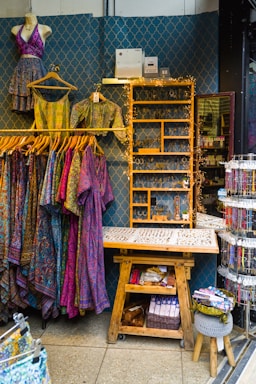 A retail display area showcasing vibrant, patterned clothing items on hangers, including dresses and tops. The wall behind has a decorative geometric pattern and a shelf adorned with fairy lights, while various accessories like jewelry are neatly organized on a wooden table. Below, there's a wooden bench with storage featuring books and a basket.