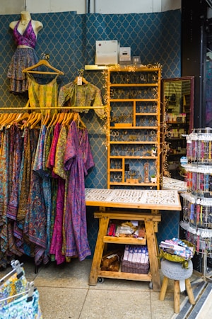 A retail display area showcasing vibrant, patterned clothing items on hangers, including dresses and tops. The wall behind has a decorative geometric pattern and a shelf adorned with fairy lights, while various accessories like jewelry are neatly organized on a wooden table. Below, there's a wooden bench with storage featuring books and a basket.