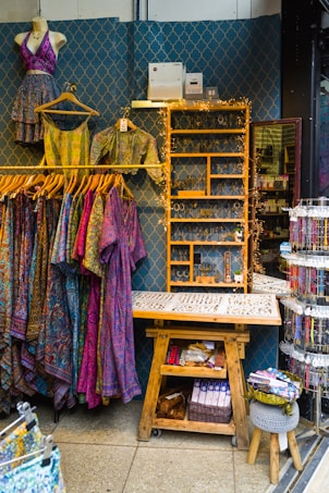 A retail display area showcasing vibrant, patterned clothing items on hangers, including dresses and tops. The wall behind has a decorative geometric pattern and a shelf adorned with fairy lights, while various accessories like jewelry are neatly organized on a wooden table. Below, there's a wooden bench with storage featuring books and a basket.