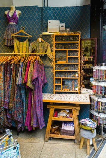 A retail display area showcasing vibrant, patterned clothing items on hangers, including dresses and tops. The wall behind has a decorative geometric pattern and a shelf adorned with fairy lights, while various accessories like jewelry are neatly organized on a wooden table. Below, there's a wooden bench with storage featuring books and a basket.