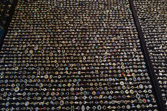 An extensive display of various rings arranged in neat rows. The rings showcase an array of gemstones in different colors and sizes, with intricate designs and settings. The background is a dark surface that highlights the sparkling jewels.