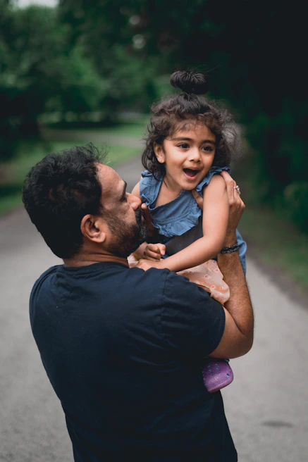 Close-up of a smiling father holding his daughter's hands as they walk together on a leafy path.