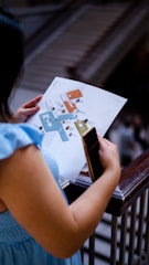 A person is holding a map or floor plan in one hand and a smartphone in the other while standing near a railing. The background appears to feature stairs or an escalator.