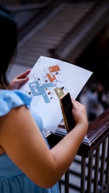 A person is holding a map or floor plan in one hand and a smartphone in the other while standing near a railing. The background appears to feature stairs or an escalator.
