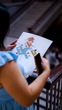 A person is holding a map or floor plan in one hand and a smartphone in the other while standing near a railing. The background appears to feature stairs or an escalator.