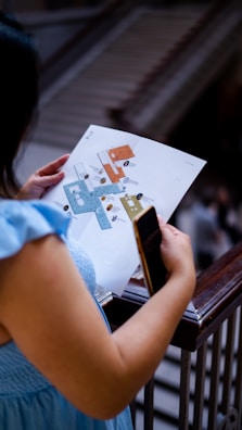 A person is holding a map or floor plan in one hand and a smartphone in the other while standing near a railing. The background appears to feature stairs or an escalator.