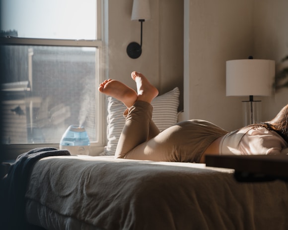 A person is lying on a bed in a cozy room with sunlight streaming through a window. Their feet are visible, and they are wearing light-colored clothing. A humidifier with visible mist sits on a table next to the window. The room is softly lit with warm tones and contains a lamp and some pillows against the wall.