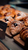 Several golden-brown pastries rest on a wooden board, each topped with powdered sugar and a dollop of dark jam. The pastries are arranged in a display setting, possibly in a bakery or cafe.