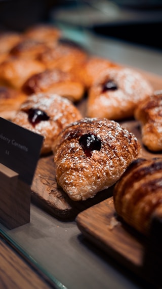 Several golden-brown pastries rest on a wooden board, each topped with powdered sugar and a dollop of dark jam. The pastries are arranged in a display setting, possibly in a bakery or cafe.