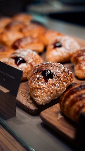 Several golden-brown pastries rest on a wooden board, each topped with powdered sugar and a dollop of dark jam. The pastries are arranged in a display setting, possibly in a bakery or cafe.