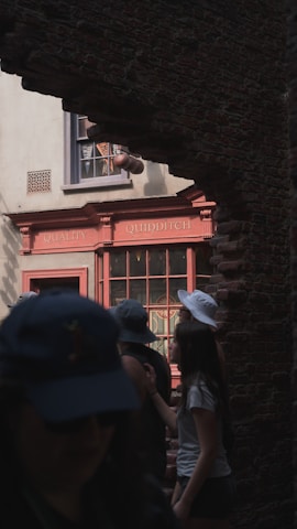 Several people are walking through an archway revealing a shop with a sign reading 'Quality Quidditch'. The scene is partially shaded, with some people wearing hats and casual clothing.