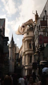 A bustling street scene featuring a large, detailed dragon perched atop an ornate building with the word 'Gringotts' displayed. The architecture is reminiscent of a fantasy world with crooked buildings and narrow alleys. A crowd of people below adds life to the scene, emphasizing the popularity and attraction of the location. The sky is partly cloudy, casting a warm light on the scene.