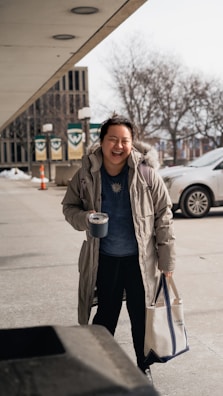 A person is standing outdoors on a sidewalk holding a reusable tote bag and a travel mug. They are wearing a heavy winter coat and appear to be smiling or laughing. Behind them, there are some banners with logos, a traffic cone, and parked cars. Leafless trees and a multi-story building are in the background.