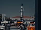 A busy urban scene with a central monument featuring a statue on a tall pillar. Below it, a fountain is surrounded by people walking and socializing. On the left, there's a white tuk-tuk vehicle, and on the right, an orange tour vehicle. Several people are captured mid-movement, and the weather appears to be sunny.