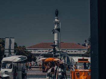 A busy urban scene with a central monument featuring a statue on a tall pillar. Below it, a fountain is surrounded by people walking and socializing. On the left, there's a white tuk-tuk vehicle, and on the right, an orange tour vehicle. Several people are captured mid-movement, and the weather appears to be sunny.
