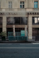Two tall, red-and-white striped ventilation pipes are positioned on a city sidewalk, enclosed by a green metal fence. Steam or smoke is rising from the pipes, partially obscuring the view of the multi-story stone building behind them. The building features large windows and an ornate architectural style. A potted plant is situated next to the enclosure. The street is empty and relatively quiet, with no visible pedestrians or vehicles.