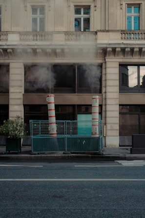 Two tall, red-and-white striped ventilation pipes are positioned on a city sidewalk, enclosed by a green metal fence. Steam or smoke is rising from the pipes, partially obscuring the view of the multi-story stone building behind them. The building features large windows and an ornate architectural style. A potted plant is situated next to the enclosure. The street is empty and relatively quiet, with no visible pedestrians or vehicles.