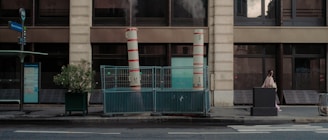 A street scene with two industrial chimneys emitting steam. The chimneys are enclosed by blue metal fencing. Nearby, there is a bus stop sign and a timetable display. A potted plant is placed next to the fence. A pedestrian wearing a mask is walking past, surrounded by an urban building facade with large windows.