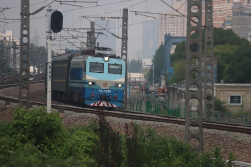 A blue and grey locomotive is traveling on a curved railway track surrounded by greenery in an urban setting. Overhead power lines and tall buildings are visible in the background. A green traffic signal and an electrical pole marked with the number 048 stand near the tracks.
