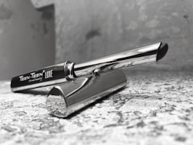 A sleek and modern pen displayed on a textured surface. The pen is silver with polished metallic parts and a black tip. The brand name 'Teen-Teen Luxe' is clearly visible on the side. The setting appears minimalist with a slightly blurred background.