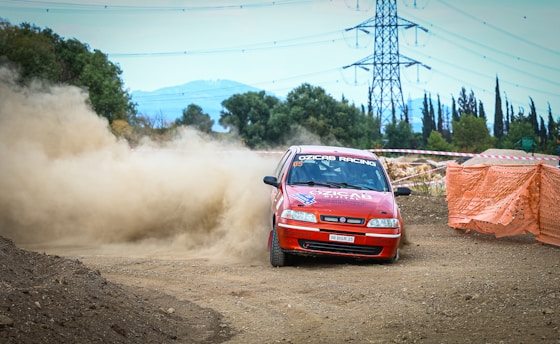 A red rally car is drifting around a dirt track corner, kicking up a large cloud of dust. It is surrounded by safety barricades, with an electrical tower and trees in the background. The car bears sponsorship decals and racing insignia.