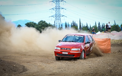 A red rally car speeds around a dirt track, kicking up a large cloud of dust behind it. The car is adorned with various sponsor logos, and the surroundings suggest an outdoor racing environment with orange barriers. Spectators and a photographer can be seen in the background, observing the event.
