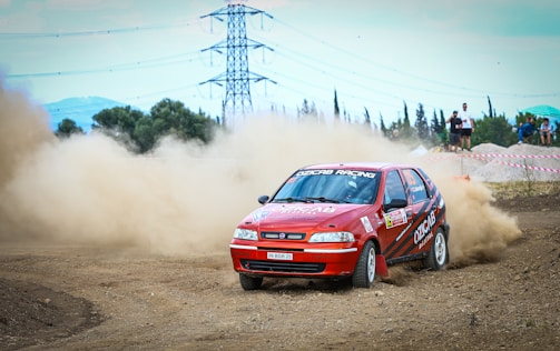 A red rally car drifts around a dirt track, kicking up a cloud of dust. The car has racing decals and sponsors on its body. In the background, there are trees, power lines, and a few spectators watching from behind a barrier.