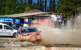 A red racing car with the label OZICAB RACING on its windshield is driving through a dusty, off-road track. There are several spectators standing nearby, observing the race. The background includes trees, a building with a red-tiled roof, and various tents and vehicles.