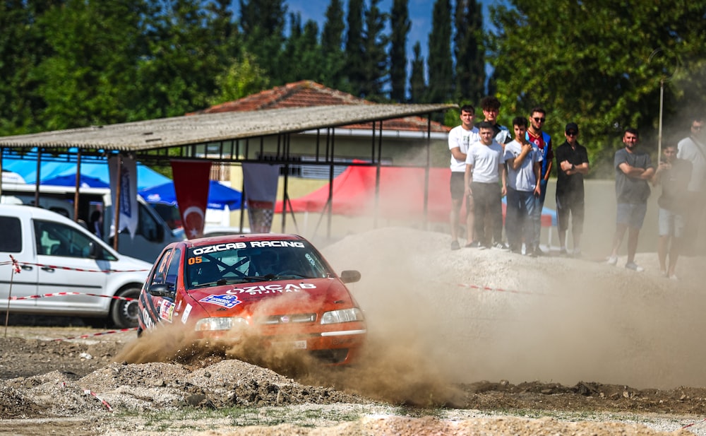 a red car driving down a dirt road next to a group of people