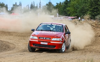 A red rally car is drifting on a dirt racing track, kicking up dust in its wake. The car has various sponsorship stickers and the words 'OZICAB RACING' on the windshield. Trees and sparse vegetation are visible in the background along with some spectators behind a barrier.