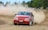 A red rally car is drifting on a dirt racing track, kicking up dust in its wake. The car has various sponsorship stickers and the words 'OZICAB RACING' on the windshield. Trees and sparse vegetation are visible in the background along with some spectators behind a barrier.