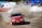A red racing car with the label 'OZICAB RACING' drifts on a dirt track, kicking up a cloud of dust. Spectators stand behind protective barriers. A white ambulance and several tents are visible in the background.