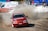 A red racing car with the label 'OZICAB RACING' drifts on a dirt track, kicking up a cloud of dust. Spectators stand behind protective barriers. A white ambulance and several tents are visible in the background.