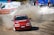 A red racing car with the label 'OZICAB RACING' drifts on a dirt track, kicking up a cloud of dust. Spectators stand behind protective barriers. A white ambulance and several tents are visible in the background.