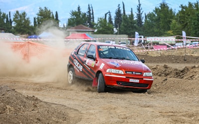 A red rally car with decals and the text 'OZCAB RACING' navigates through a dirt track, kicking up dust as it takes a sharp turn. The background includes trees and a few structures, with red and white tape marking the track barriers.