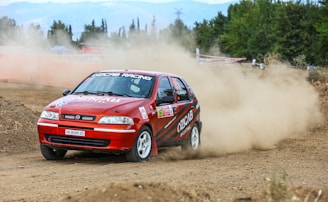 A red rally car is driving swiftly on a dirt track, kicking up a large cloud of dust behind it. The car is decorated with various sponsor logos and is navigating a curve with visible speed and agility. In the background, there are green trees and some mountainous terrain, under a cloudy sky.