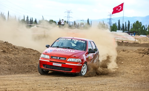 A red racing car drifts around a dirt track, kicking up a substantial cloud of dust. The vehicle displays various racing decals and a visible number plate. A large flag can be seen in the background, along with some trees and power lines under a partly cloudy sky.