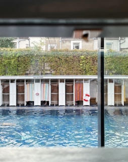 A view of a swimming pool surrounded by cabana-like structures with colorful curtains. Above the cabanas, a sign reads 'No Diving.' Behind the cabanas is a wall covered with green ivy. The pool water is a vibrant blue, reflecting light.
