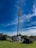 A tall communication tower surrounded by greenery under a vibrant blue sky with scattered clouds. The fenced area at the base contains technical equipment.